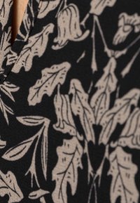 Svart tyg med ett krämfärgat blommönster som innehåller blad och blommor. Texturen är slät med en svag glans. Noterbar v-ringning.