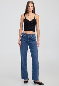 Žena stojící v černém bezrukávovém crop topu s třpytkami, v modrých džínech s vysokým pasem a rovnými nohavicemi, a v černých balerínkách na světlém pozadí.