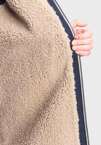Beige fleecetextiel met een getextureerd oppervlak, gecombineerd met een marineblauwe rits en afwerking. Een hand houdt de rand van het materiaal vast.