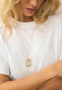 Hängande halsband i guldton med sammankopplade ovala former, hängande från ett tunt kedja, bärs över en vit t-shirt med korta ärmar.