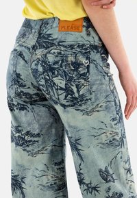 Pantalones de mezclilla anchos de color azul claro con patrones florales oscuros, que presentan una etiqueta de cinturón de cuero marrón con "PLEASE" y un acento en forma de corazón en el bolsillo.