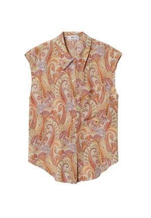 Kobieta w bezrękawowej bluzce z wzorem paisley, jasnych prostych spodniach i czarnych sandałach na blokowym obcasie, stojąca.