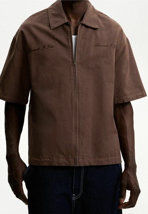 Mężczyzna w brązowej koszuli na krótki rękaw z zamkiem i kołnierzykiem oraz ciemnych spodniach, stojący z rękami wzdłuż ciała na gładkim tle.