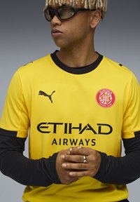Gul sports trøje med sorte accenter, der har Puma-logoet, teksten "ETIHAD AIRWAYS" og Girona FC-kresten. Sorte langærmede trøjer ses underneden.