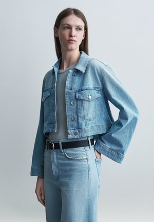 Młoda kobieta w jasnoniebieskiej kurtce jeansowej i dżinsach, szarym swetrze i czarnym pasku, z ręką w kieszeni, patrząca na bok.