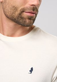 T-Shirt od kremaste pamučne tkanine s okruglim izrezom i malim plavim vezom na lijevoj strani prsa. Mekana tekstura, jednostavan dizajn.