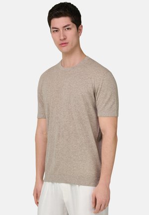 Uomo in piedi che indossa una maglia a maniche corte beige lavorata a maglia e pantaloni bianchi su uno sfondo chiaro e uniforme.