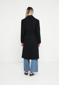 Cappotto di lana nero con cintura, maniche lunghe e colletto strutturato, abbinato a jeans blu a gamba larga e tacchi neri.