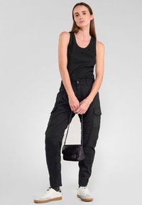 Zwarte tanktop, cargobroek met zijzakken en witte sneakers. Kleine zwarte handtas met kettingband. Gladde stof en aansluitend ontwerp.