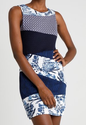 Femme portant une robe sans manches, ajustée, longueur genou avec des motifs floraux et géométriques bleus et blancs, main reposant sur la hanche.