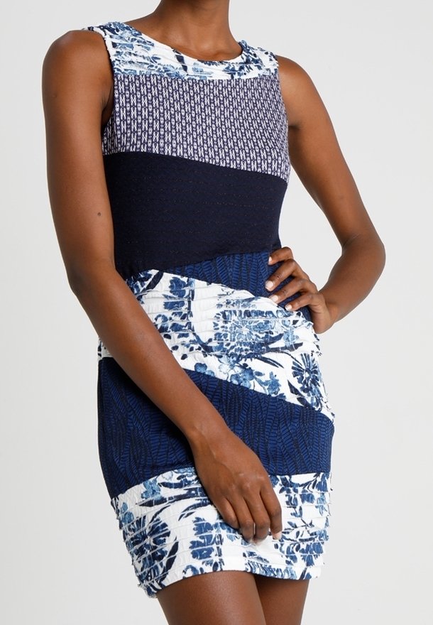 Femme portant une robe sans manches, ajustée, longueur genou avec des motifs floraux et géométriques bleus et blancs, main reposant sur la hanche.
