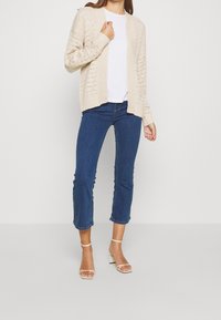 Beige stickad kappa med öppen front, matchad med en vit t-shirt och blå cropped jeans. Klara blockklacksandaler fulländar looken.