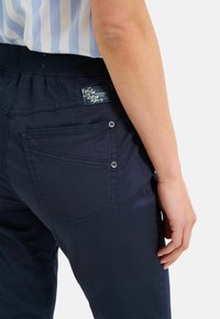 Námořnické modré kalhoty s jemným leskem, vybavené dvěma zadními kapsami, štítkem s logem a patentními uzávěry na kapsách, vyrobené z lehkého materiálu.