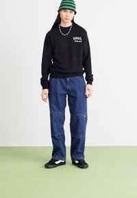 Black sweatshirt with "DICKIES" logo, blue denim cargo pants, and black sneakers with white accents. Model wears a green-striped beanie.
