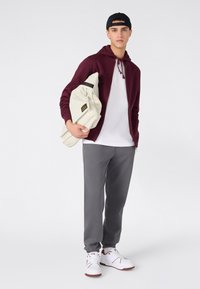 Jonge man in een zwarte pet, bordeaux rood hoodie met rits, wit T-shirt, grijze joggers en witte sneakers, die een opgevouwen beige jas vasthoudt.