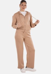 Felpa beige con zip e coulisse abbinata a pantaloni della tuta beige, entrambi realizzati in tessuto morbido. Sneakers bianche completano l'outfit.