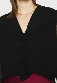 Blusa nera in tessuto testurizzato, con colletto arricciato, chiusura anteriore con bottoni e maniche trasparenti, abbinata a una collana di catena dorata.