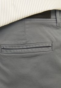 Gros plan sur la poche arrière d'un pantalon gris avec une ouverture passepoilée et une chemise crème à fines rayures rentrée à l'intérieur.