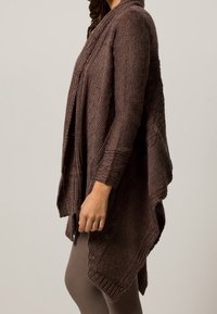 Dziergany sweter rozpinany w ciemnym brązie z drapowanym krojem. Charakteryzuje się teksturowaną tkaniną i dłuższym tylnym brzegiem, noszony na dopasowanym topie i legginsach.