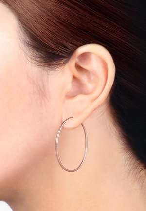 Roséfarben, große Creole mit einer glatten, reflektierenden Oberfläche. Dünnes Drahtdesign, das am Ohr gegen dunkles Haar präsentiert wird.