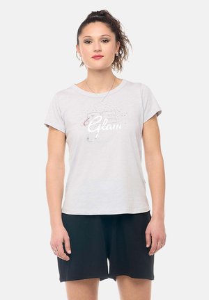 Jeune femme aux cheveux attachés, portant un t-shirt blanc « Glam » et un short noir, debout devant un fond clair uni.