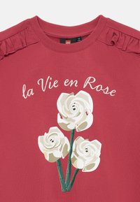 T-shirt in cotone rosso con graphic di rose bianche e testo "la Vie en Rose". Gli accenti arruffati sulle spalle aggiungono un dettaglio di texture.