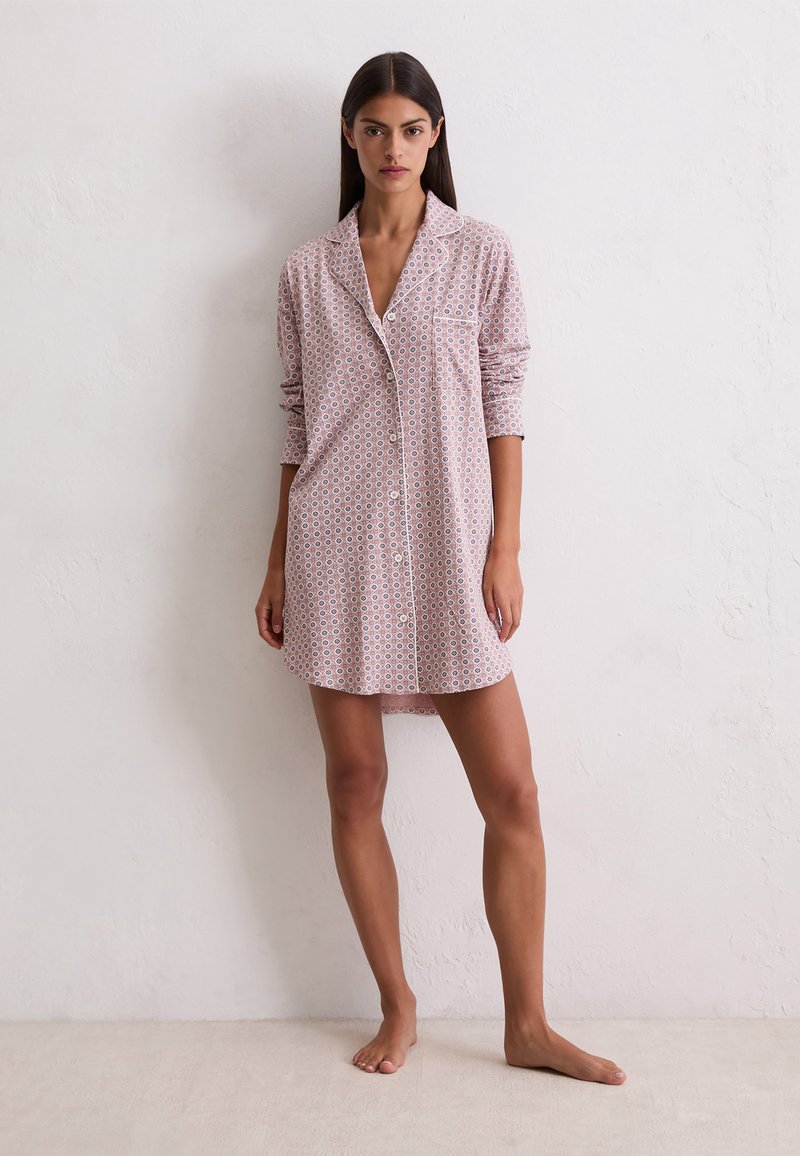 Femme debout pieds nus contre un mur portant une chemise de nuit rose à manches longues et à boutons avec un petit motif géométrique.