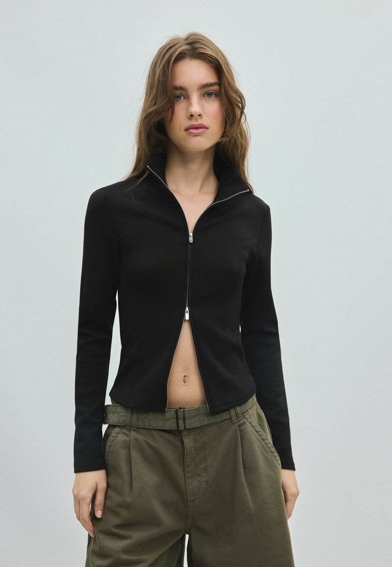 Jeune femme aux longs cheveux bruns porte un haut noir zippé partiellement ouvert et un pantalon ample vert olive sur un fond clair uni.