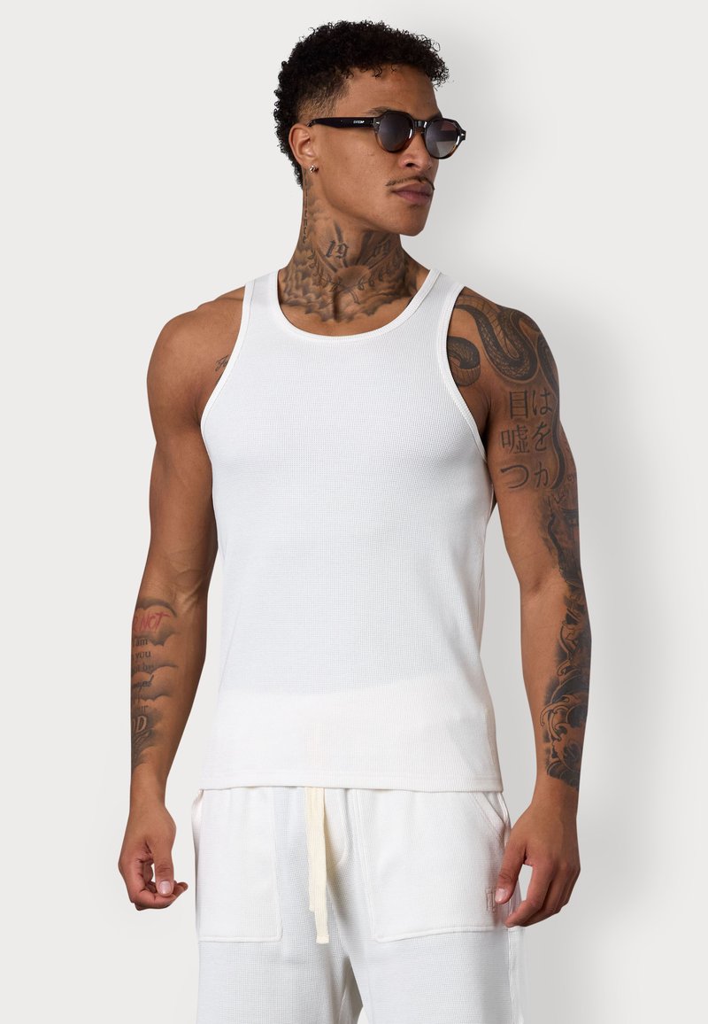 Weißes ärmelloses Tanktop aus geripptem Stoff, mit Rundhalsausschnitt und figurnaher Passform, kombiniert mit hellen Jogginghosen mit Kordelzug.