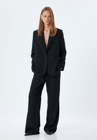 Zwarte op maat gemaakte blazer met een diepe V-hals, gecombineerd met wijde broekspijpen. Glad materiaal, subtiele zakken en minimalistische ontwerpdetails.