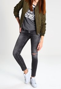 Olivgrüne Bomberjacke, graues T-Shirt mit Grafik, schwarze Skinny Jeans mit Knie-Rissen und weiße Sneakers. Einfache, urbane Outfit-Zusammenstellung.