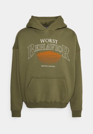 Hoodie vert olive avec cordon de serrage, poche frontale, et texte imprimé en blanc et orange : « PIRE COMPORTEMENT » avec des lignes horizontales.
