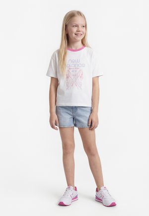 Niña joven de pie, vestida con camiseta blanca de New Balance con gráfico de mariposa, pantalones cortos de mezclilla azul claro y zapatillas blancas con rosa.