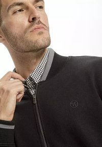 Czarna bomberka zapinana na zamek błyskawiczny z prążkowanym kołnierzykiem i mankietami, z szarymi paskami. Pod spodem widoczna jest czarno-biała koszula w kratę.