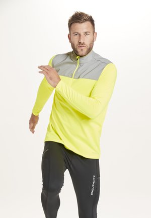 Chemise de sport à manches longues de couleur jaune vif avec un panneau gris et un col zippé, associée à des leggings noirs avec une poche latérale et un logo.