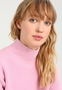 Sølvstjernede øreringe udstillet mod en lys baggrund, med en poleret overflade og detaljeret design, båret med en lyserød ribbet sweater.