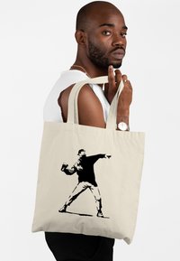 Borsa a tracolla in tela beige con silhouette nera di un uomo che lancia una bomboletta spray, dotata di manici resistenti. Design semplice e funzionale.