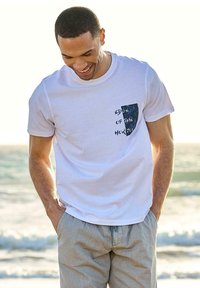 White cotton t-shirt with a blue patterned pocket featuring the text "EDGE OF THE HORIZON." Model wearing light gray shorts.