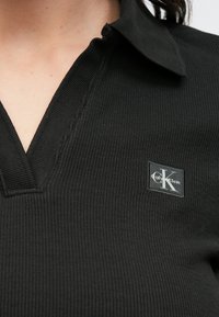 Polo nera a coste con colletto, con una piccola patch con il logo Calvin Klein sul lato sinistro. Tessuto semplice e strutturato.