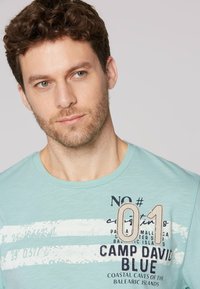 Lichtblauw katoenen T-shirt met een ronde hals, korte mouwen en bedrukte tekst met een grafisch ontwerp in donkerdere kleuren.