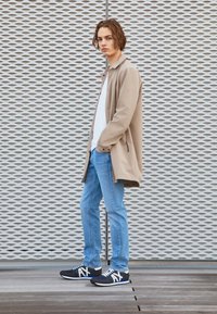 Beige lett jakke over en hvit T-skjorte, paret med lyseblå jeans og svarte joggesko med hvite logoer. Teksturert bakgrunn med mønstre.