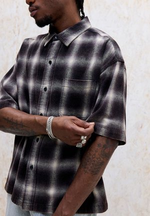 Homme remontant la manche d'une chemise à manches courtes à carreaux noirs et blancs, montrant des tatouages sur les bras et portant des bagues et bracelets en argent.