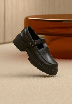 Schwarzer Schuh mit grober Lederlaufsohle und Schnallenriemen, schwebend über einem beigen Teppich neben einem braunen ovalen Tisch dargestellt.