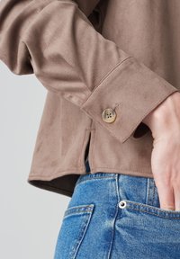 Brown suede shirt with a buttoned cuff, featuring a soft texture and side slits. Paired with blue denim jeans.