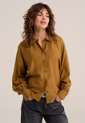 Femme aux cheveux bouclés portant un chemisier moutarde à manches longues et un pantalon noir, les mains dans les poches.