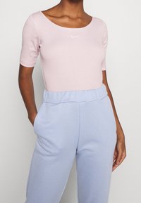 Vrouw in een lichtroze Nike t-shirt met korte mouwen, gestopt in hoogglans lavendel joggingbroek met handen in de zakken.