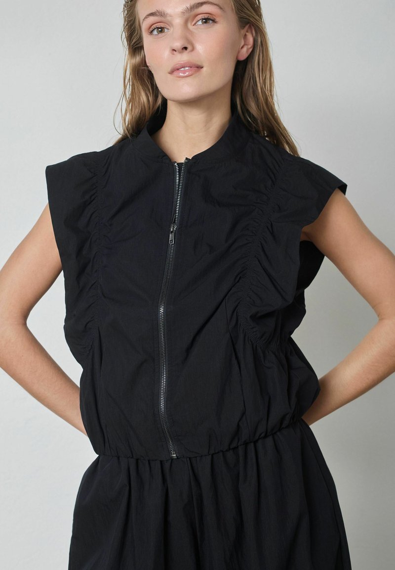 Čierne šaty bez rukávov s vysokým golierom, zapínaním na zips a nariasenými detailmi pozdĺž strán, so voľným strihom a mäkkou textúrou.