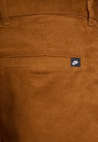 Ruskeat corduroy-housut, joissa on pystysuorat ribbinauhat ja teksturoitu pinta, takatasku, jossa pieni musta Nike-logo.