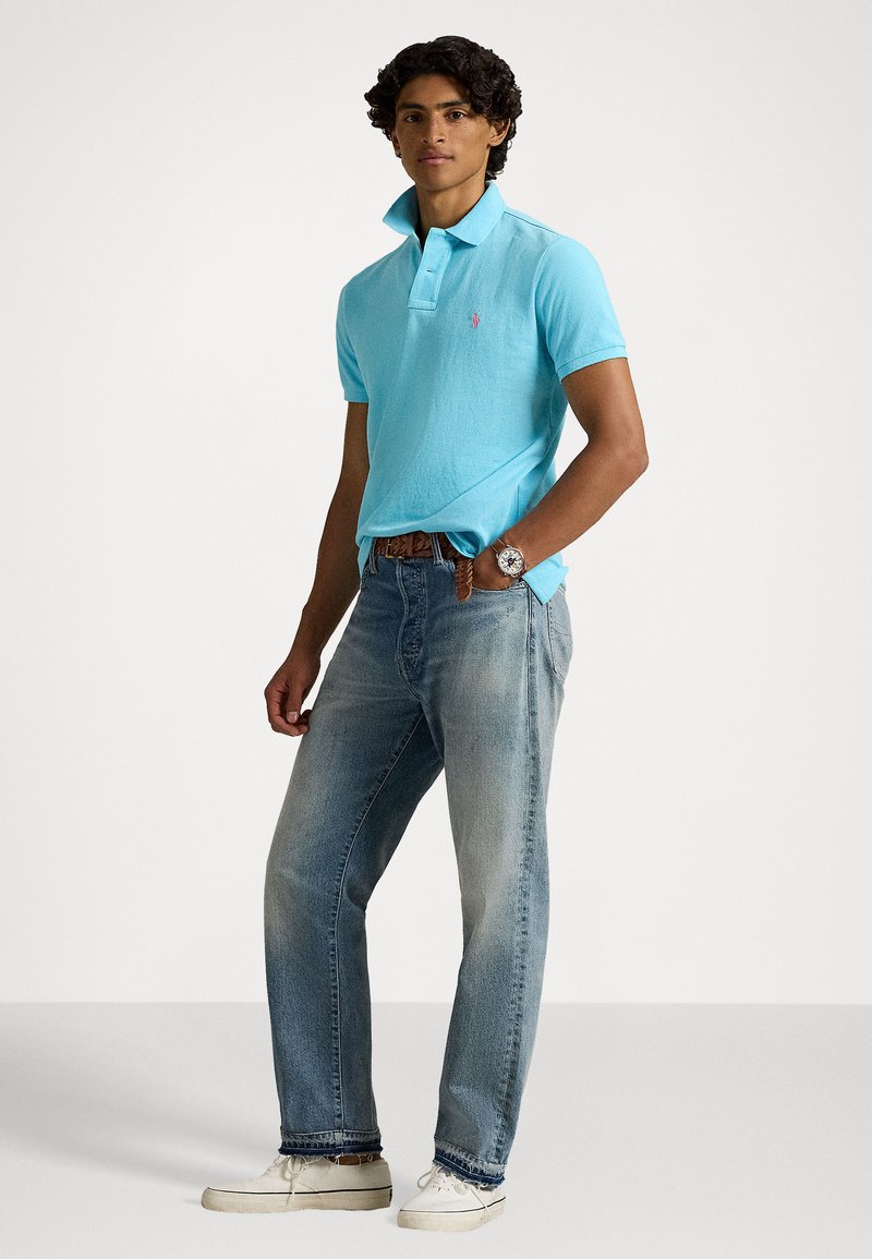 Kurzärmeliges Polohemd aus hellblauem Baumwollstoff, kombiniert mit ausgewaschenen blauen Jeans. Mit Kragen, Knöpfen und einem Logo-Akzent auf der Brust.