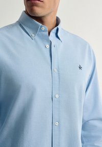 Camisa de botón azul claro hecha de algodón. Presenta un cuello con solapa, botones blancos y un pequeño logo negro en el pecho. Textura suave.